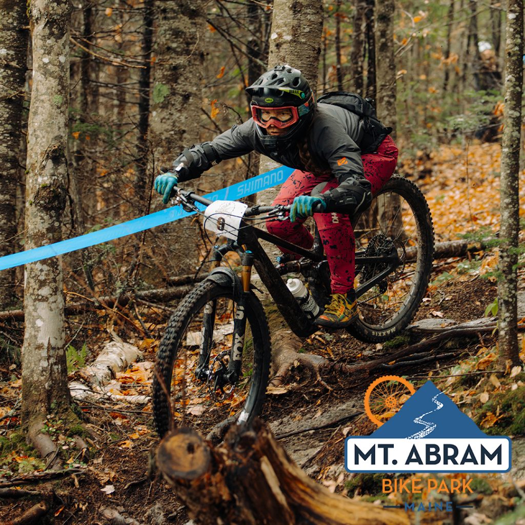 Mountain biker in a helmet and goggles navigating a rocky forest trail, number plate on handlebars, blue Shimano branding visible, Mt. Abram Bike Park logo bottom right