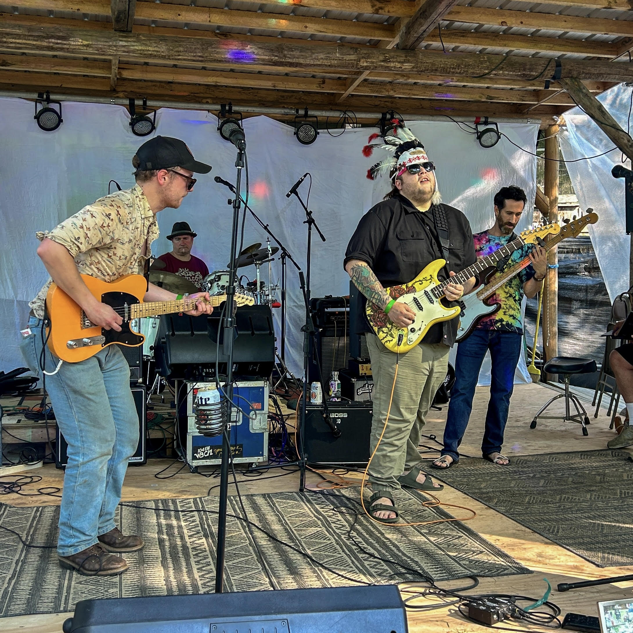 Three male musicians performing on an outdoor stage with guitars and drums, wearing casual shirts and hats under a wooden shelter.